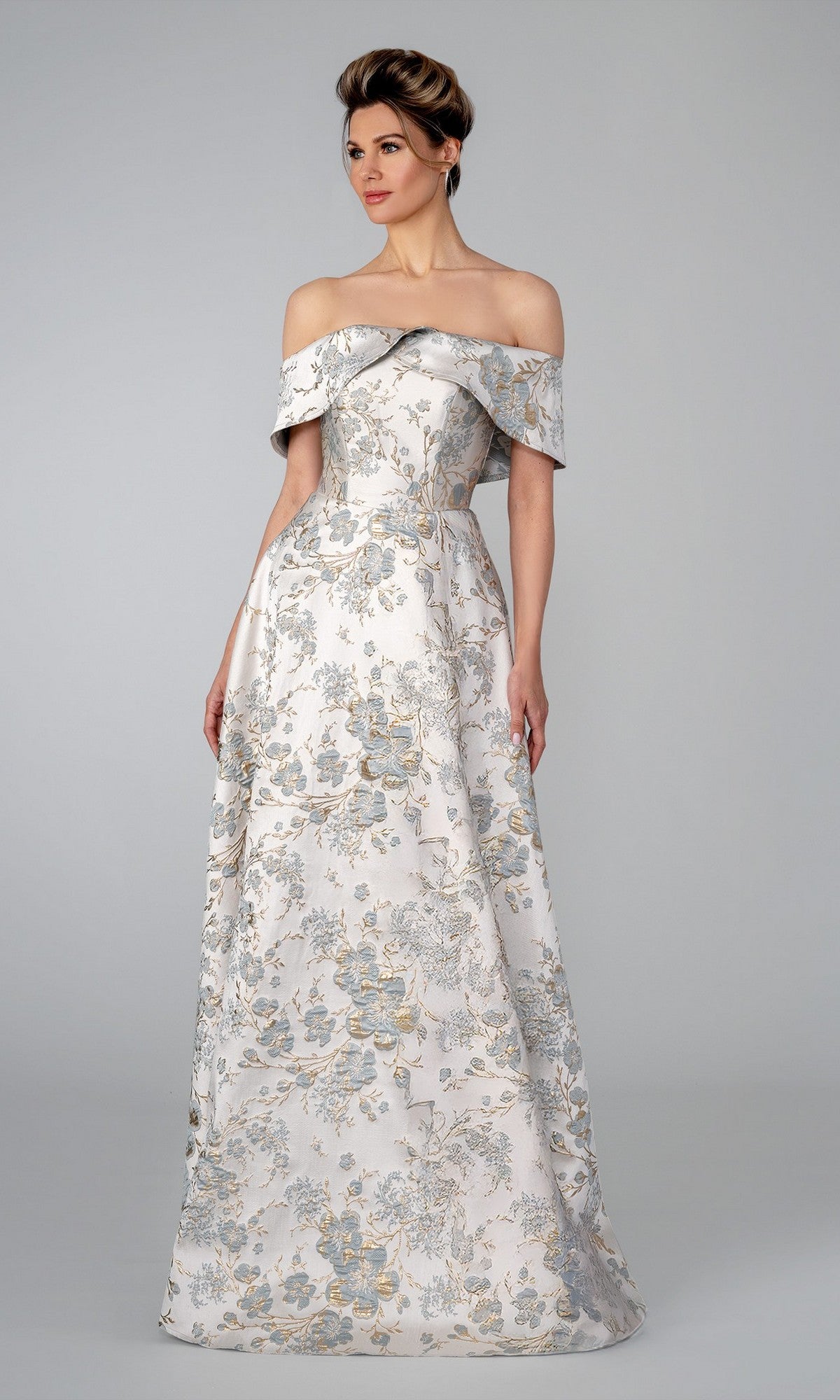 A woman in a blue Stella Couture 25303 long formal dress featuring an off-the-shoulder neckline and silver floral jacquard stands before a plain grey background.